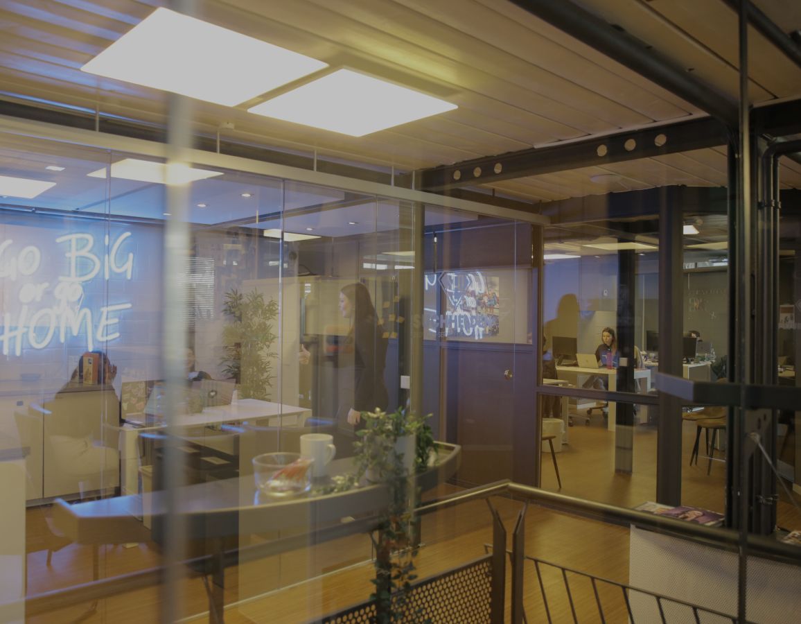 Blick durch Glaswände in ein modernes Büro mit Besprechungsraum und arbeitenden Personen im Hintergrund.