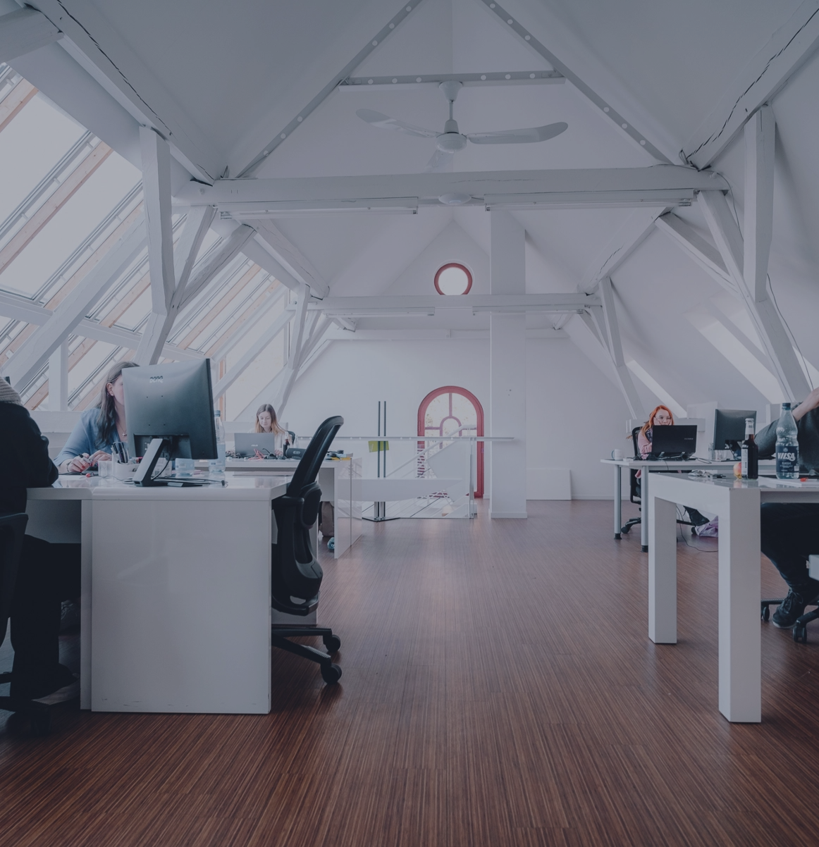 Helles Großraumbüro mit mehreren Personen an Schreibtischen unter einem Dach mit großen Fenstern.