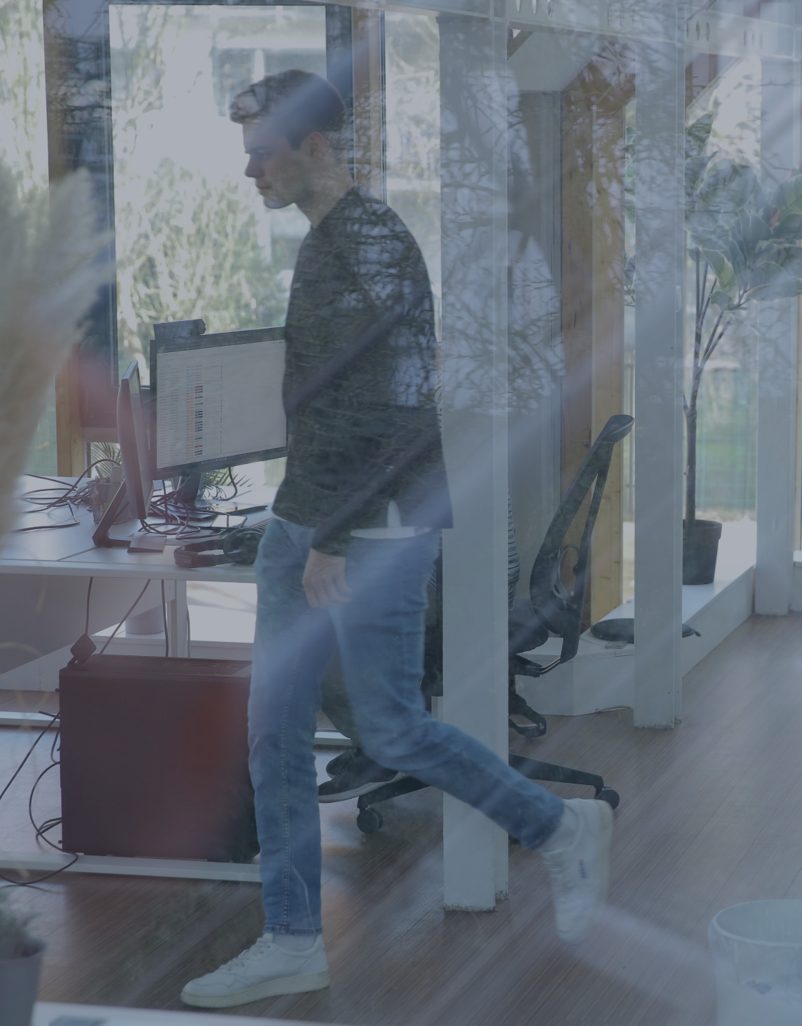 Mitarbeiter geht durch ein modernes Büro an einem Computerarbeitsplatz vorbei, aufgenommen durch eine Glaswand.