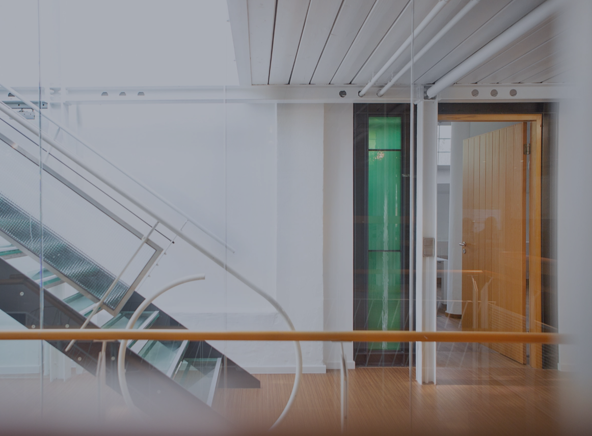 Heller Büroflur mit moderner Glas- und Metalltreppe, weißen Wänden und Holzboden; rechts ein offener Raum mit Holztür und grünem Glaselement, klare, minimalistische Architektur