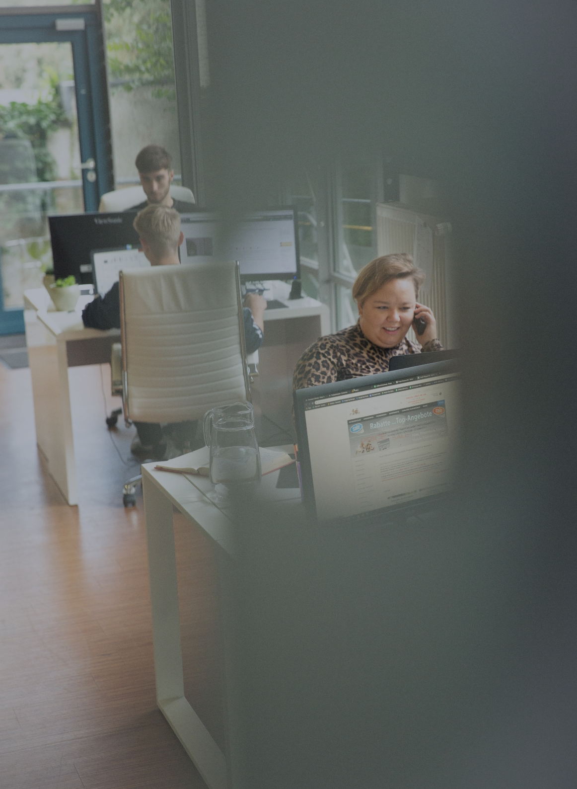 Mitarbeiterin telefoniert an ihrem Schreibtisch in einem Büro, während im Hintergrund weitere Mitarbeitende an Computerarbeitsplätzen arbeiten.