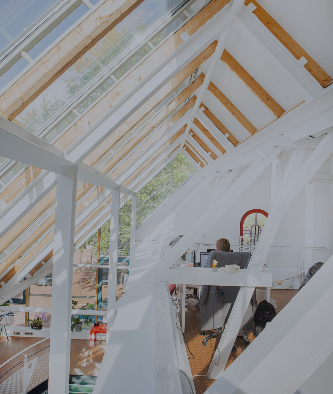 Helles Dachgeschossbüro mit großen Glasdachfenstern und sichtbaren Holz- und Stahlträgern; eine Person sitzt an einem Schreibtisch, viel Tageslicht fällt in den offenen, modernen Arbeitsbereich