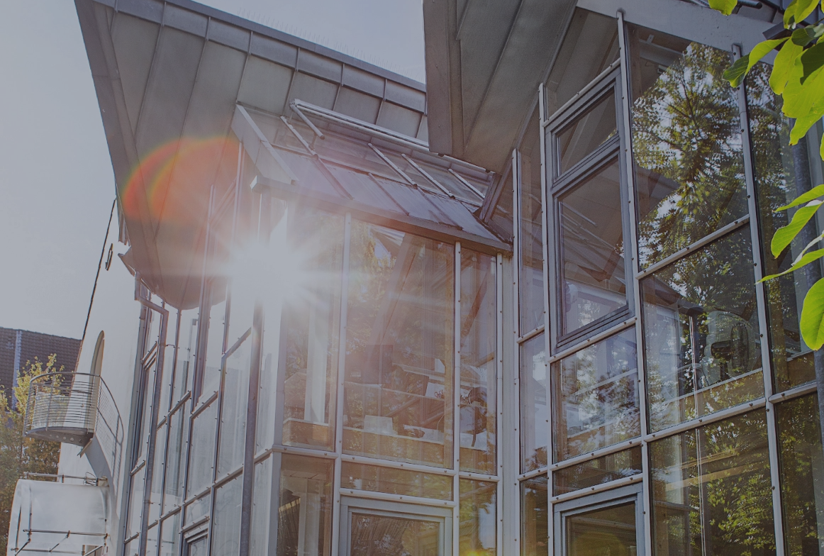 Außenansicht eines modernen Bürogebäudes mit großer Glasfassade, schrägen Dächern und Bäumen, durch die Sonnenlicht fällt