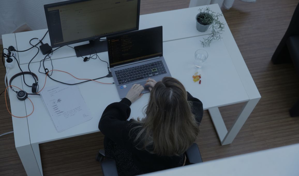 Person tippt an einem Laptop am Schreibtisch im Büro, aufgenommen aus der Vogelperspektive, mit externem Monitor und kleiner Pflanze