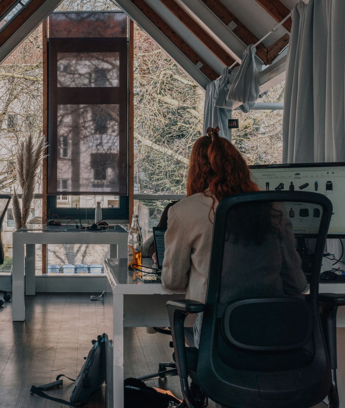 Eine Person arbeitet an einem Schreibtischen in einem hellen Büro mit großen Fenstern, sichtbaren Holzbalken und Blick ins Grüne