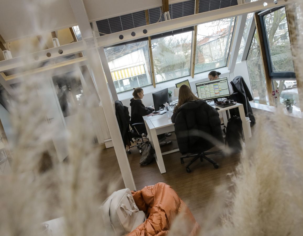 Person arbeitet an einem Schreibtisch im Büro, seitlich durch eine Glaswand fotografiert, mit Fensterfront im Hintergrund.