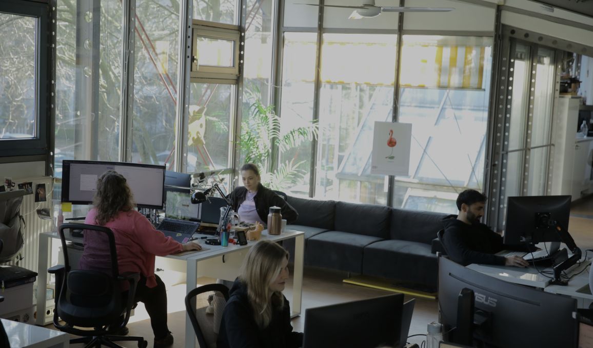Mehrere Personen arbeiten an Computern in einem hellen, modernen Büro mit großen Fenstern, Pflanzen und Sitzbereich.