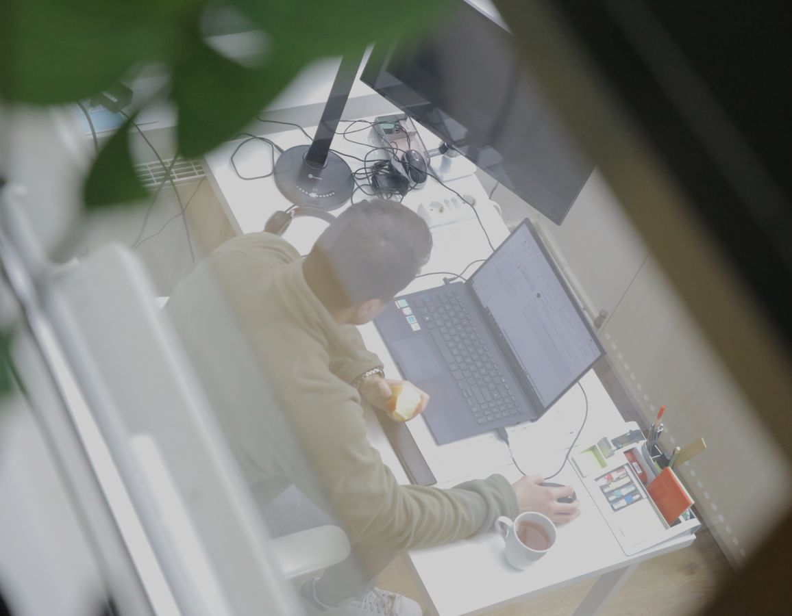 Person arbeitet an einem Laptop am Schreibtisch im Büro, hält einen Apfel in der Hand, aufgenommen aus erhöhter Perspektive