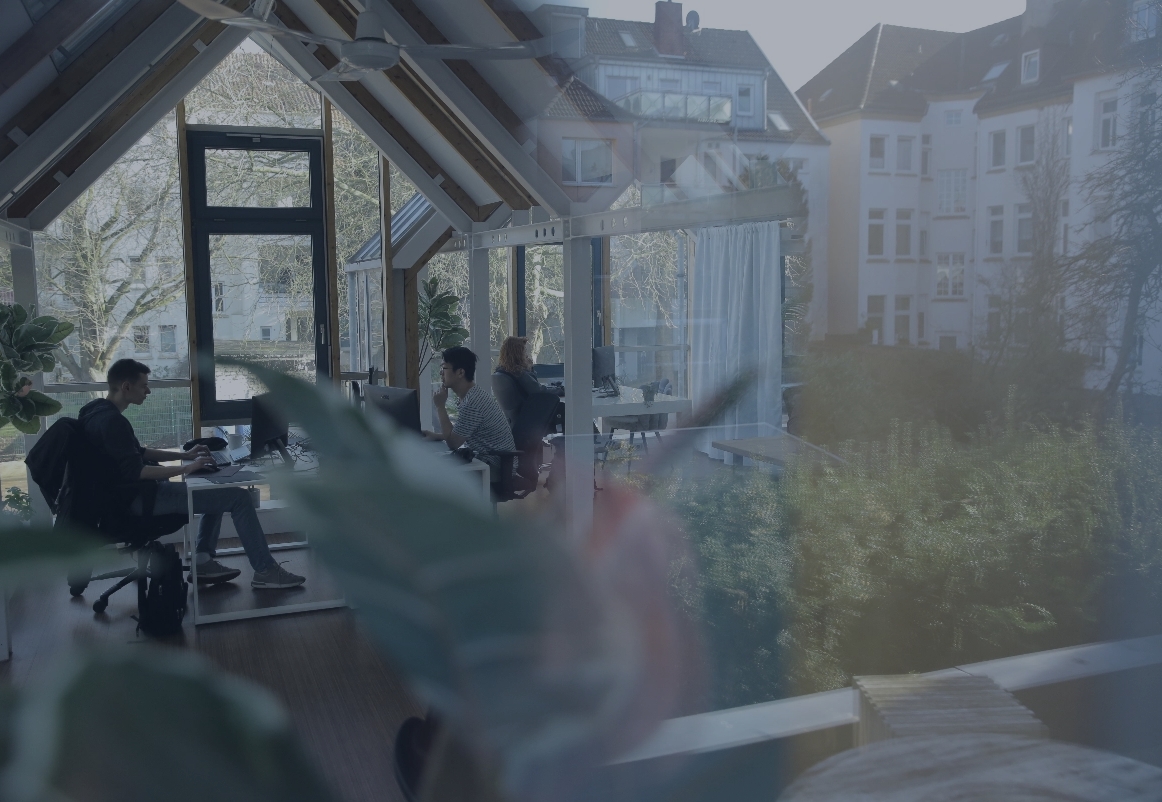 Zwei Personen arbeiten an Computern in einem hellen Büro mit großen Fenstern, Holzbalken und Pflanzen im Vordergrund.