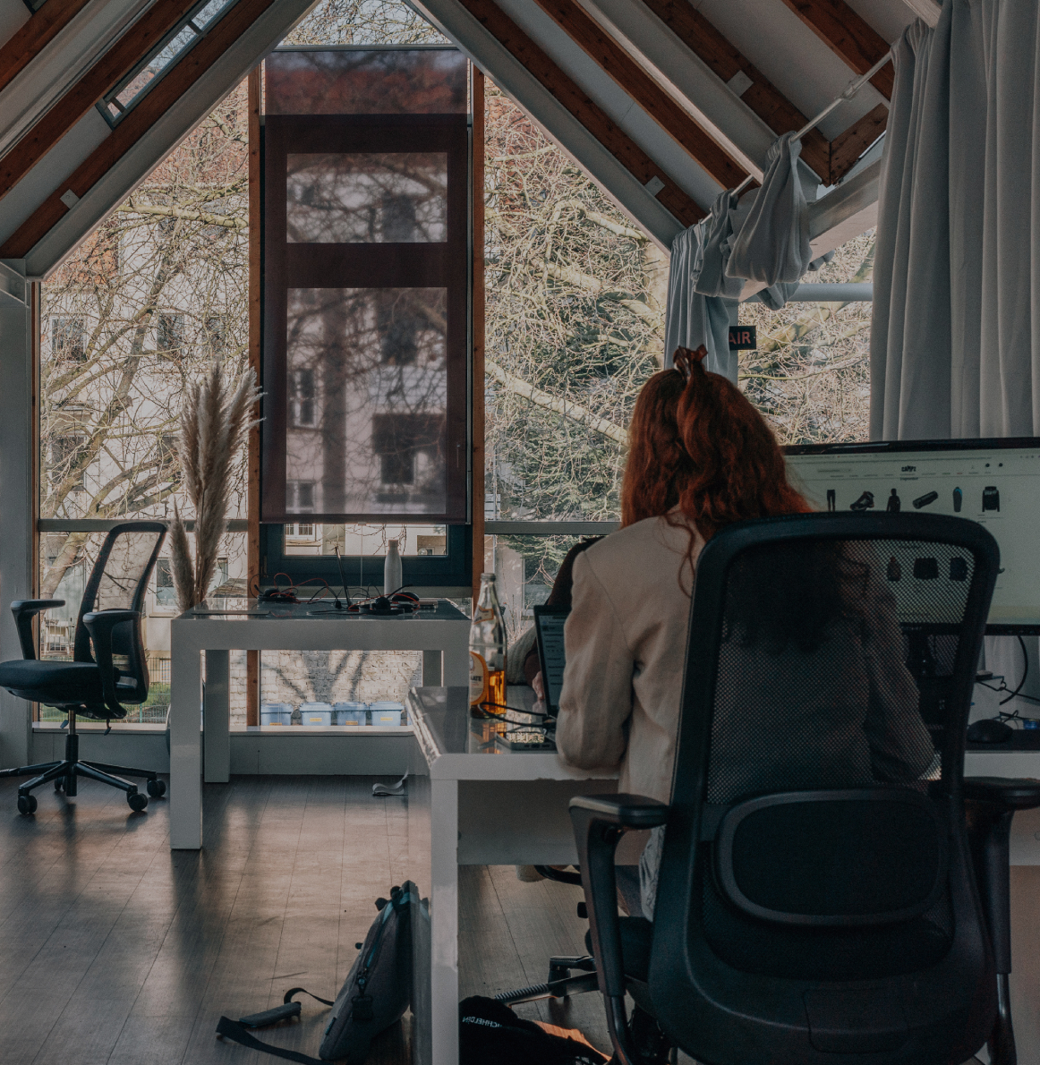 Zwei Personen arbeiten an separaten Schreibtischen in einem hellen Büro mit großen Fenstern, sichtbaren Holzbalken und Blick ins Grüne