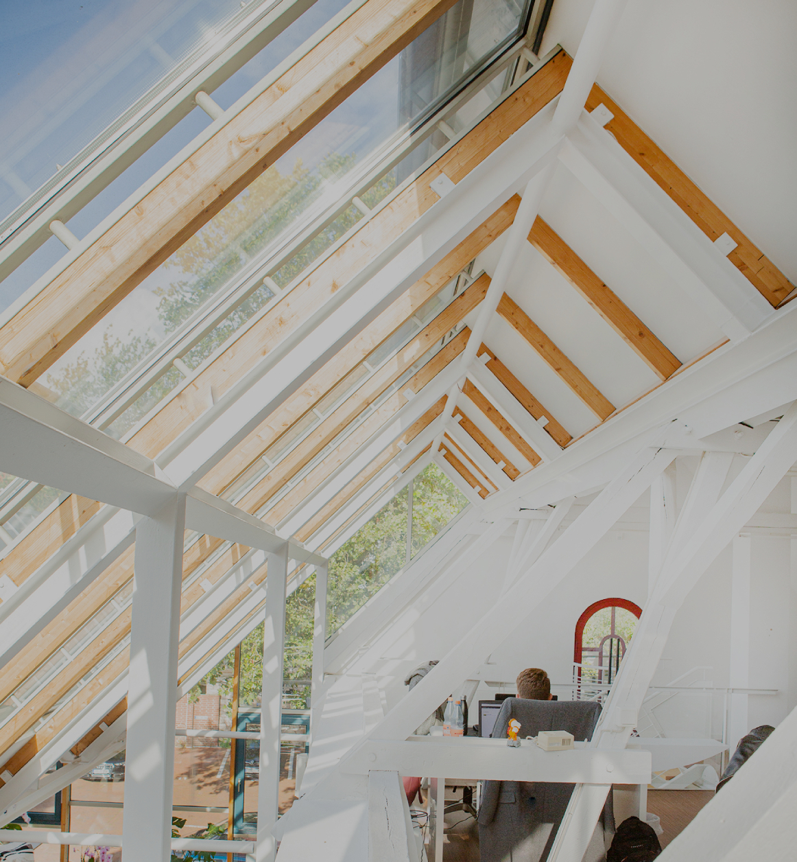 Helles Dachgeschossbüro mit großen Glasdachfenstern und sichtbaren Holz- und Stahlträgern; eine Person sitzt an einem Schreibtisch, viel Tageslicht fällt in den offenen, modernen Arbeitsbereich