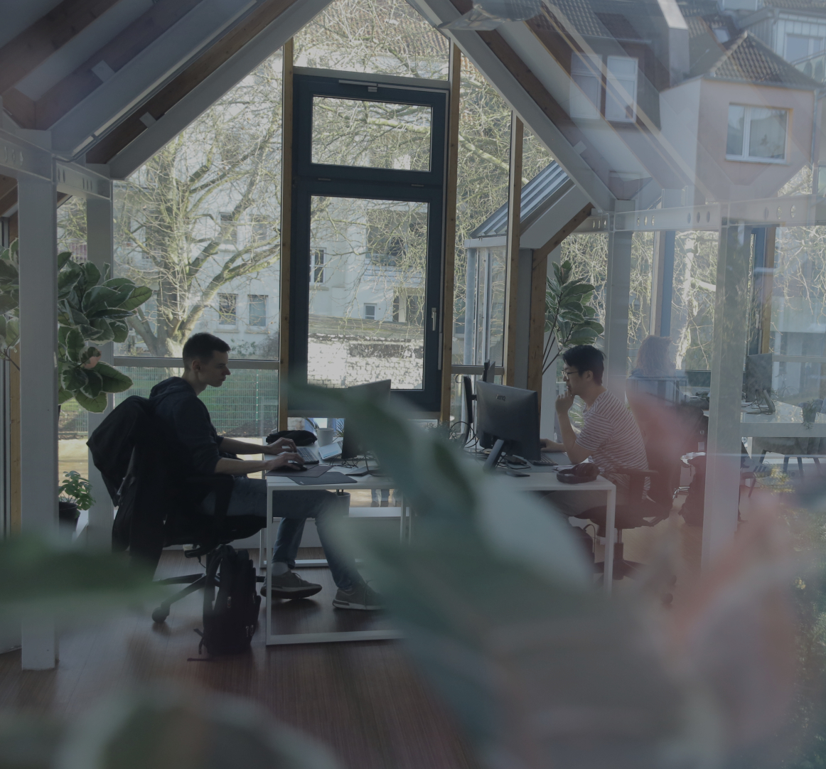 Zwei Personen arbeiten an Computern in einem hellen Büro mit großen Fenstern, Holzbalken und Pflanzen im Vordergrund.