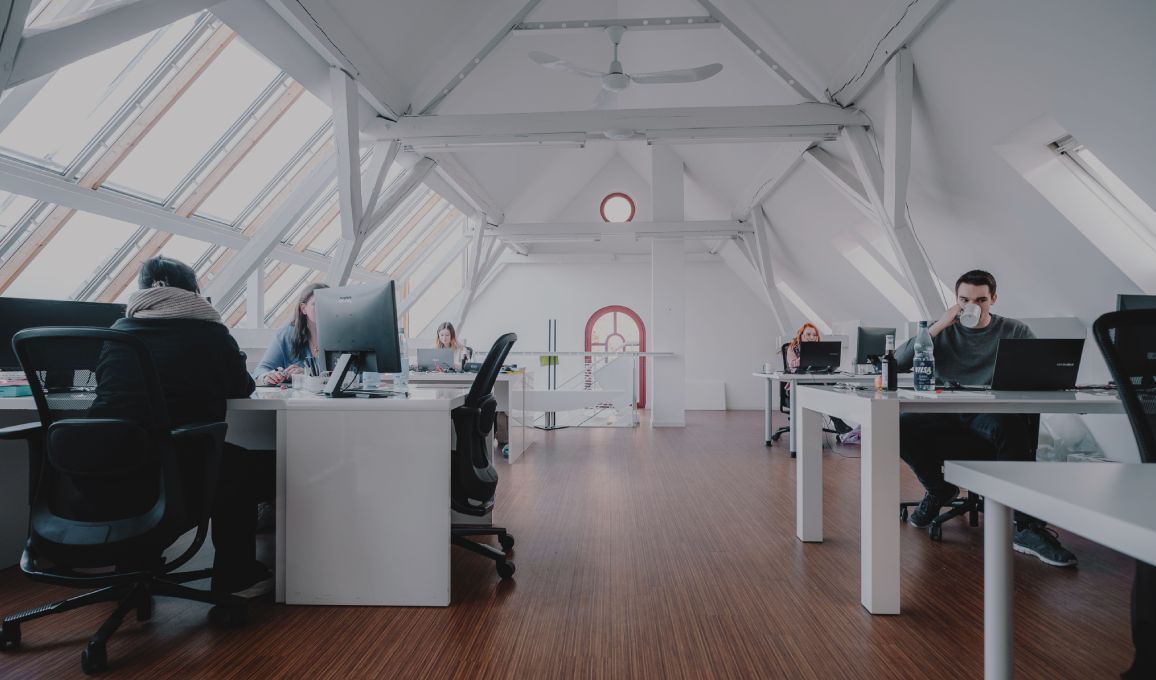 Helles Großraumbüro mit mehreren Personen an Schreibtischen unter einem Dach mit großen Fenstern.