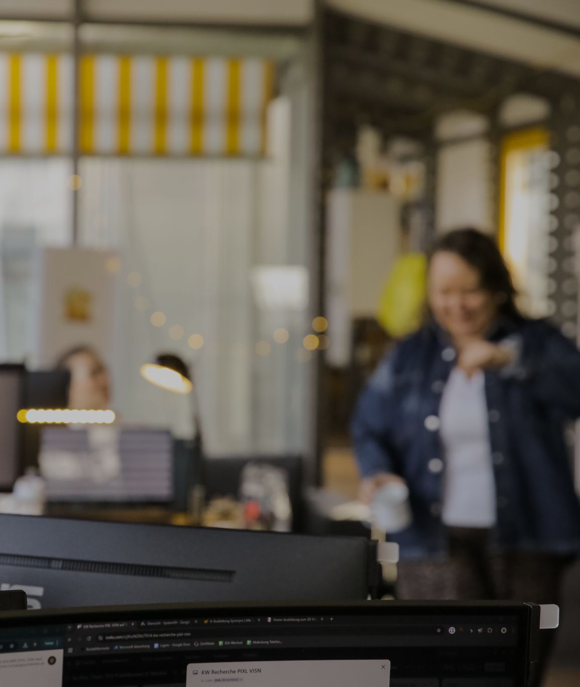 Unscharfe Büroszene mit Monitor im Vordergrund und einer Person, die im Hintergrund mit einer Tasse durch das Büro geht.