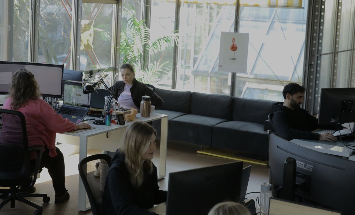 Mehrere Personen arbeiten an Computern in einem hellen, modernen Büro mit großen Fenstern, Pflanzen und Sitzbereich.