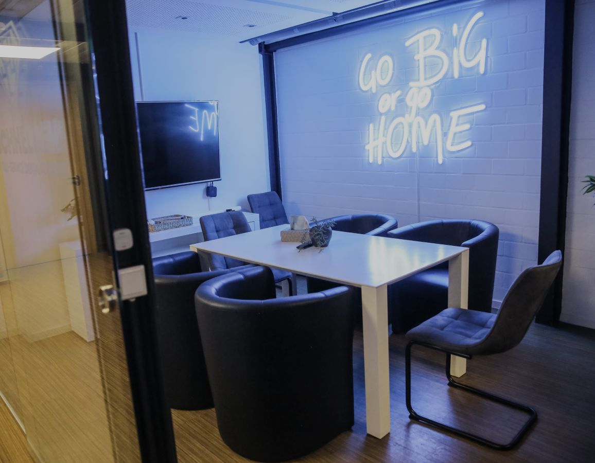 Moderner Besprechungsraum mit Tisch, Stühlen und leuchtender Neon-Schrift „Go Big or Go Home“ an der Wand.