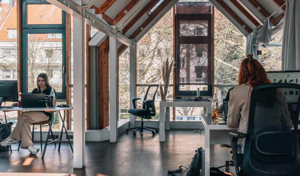 Zwei Personen arbeiten an separaten Schreibtischen in einem hellen Büro mit großen Fenstern, sichtbaren Holzbalken und Blick ins Grüne