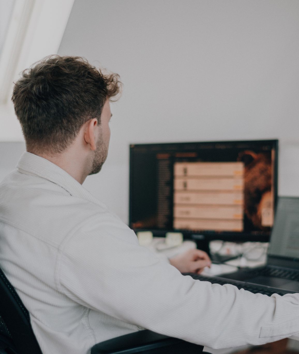 Person sitzt an einem Schreibtisch und arbeitet an einem Computer mit großem Monitor in einem hellen Büro, von hinten bzw. seitlich fotografiert