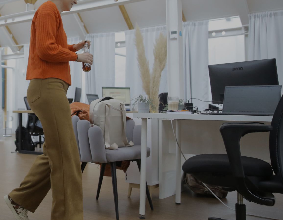 Person geht durch ein helles Büro mit Schreibtischen und Computern und hält eine Trinkflasche in der Hand.