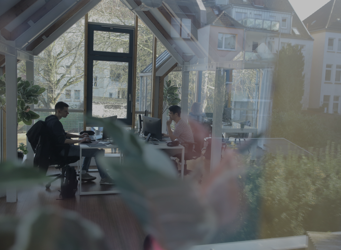 Zwei Personen arbeiten an Computern in einem hellen Büro mit großen Fenstern, Holzbalken und Pflanzen im Vordergrund.
