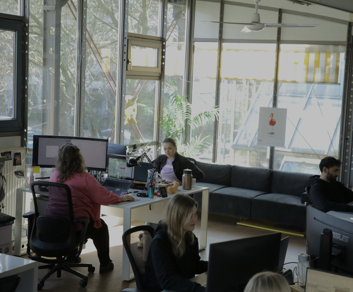 Mehrere Personen arbeiten an Computern in einem hellen, modernen Büro mit großen Fenstern, Pflanzen und Sitzbereich.