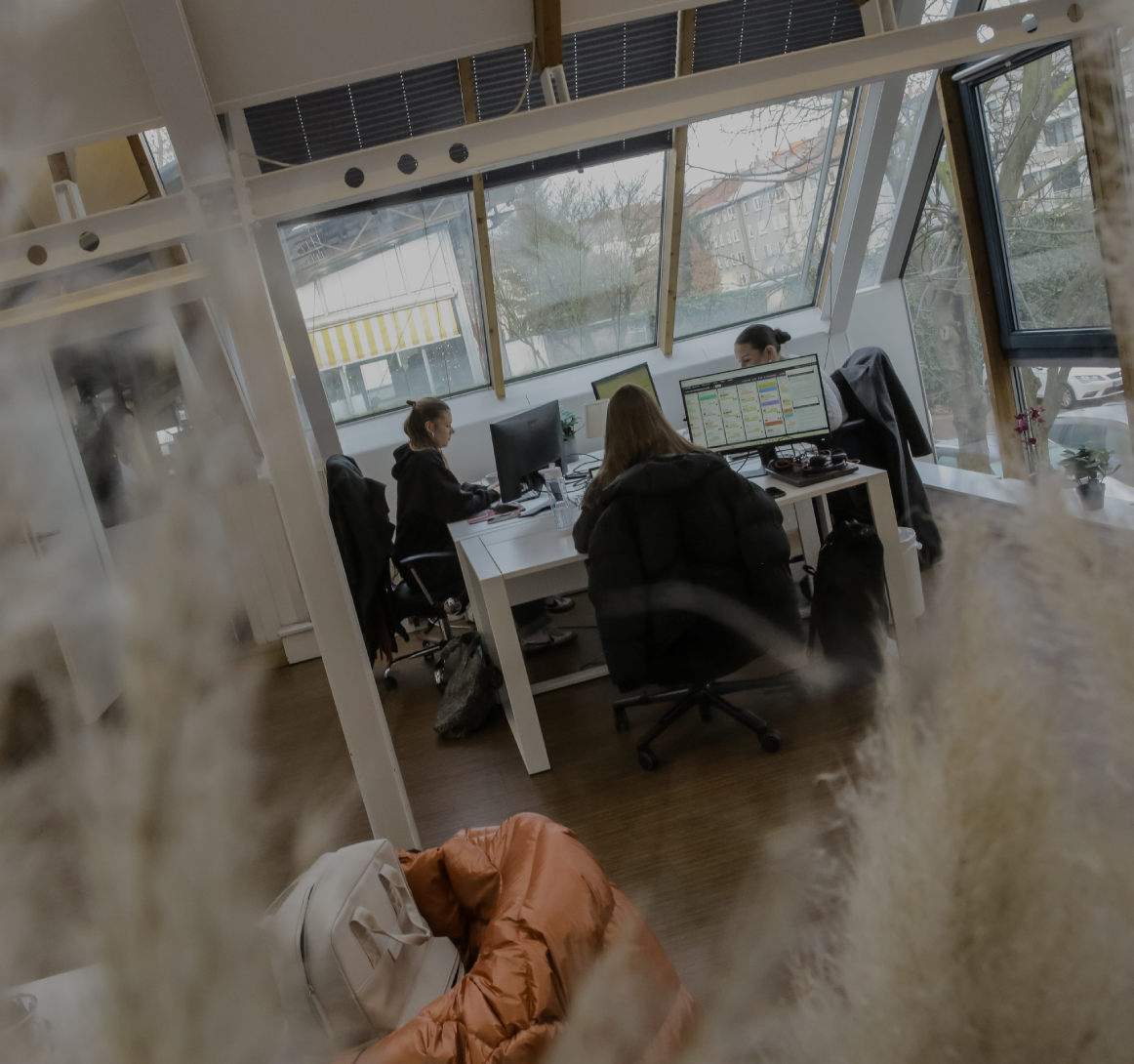 Person arbeitet an einem Schreibtisch im Büro, seitlich durch eine Glaswand fotografiert, mit Fensterfront im Hintergrund.