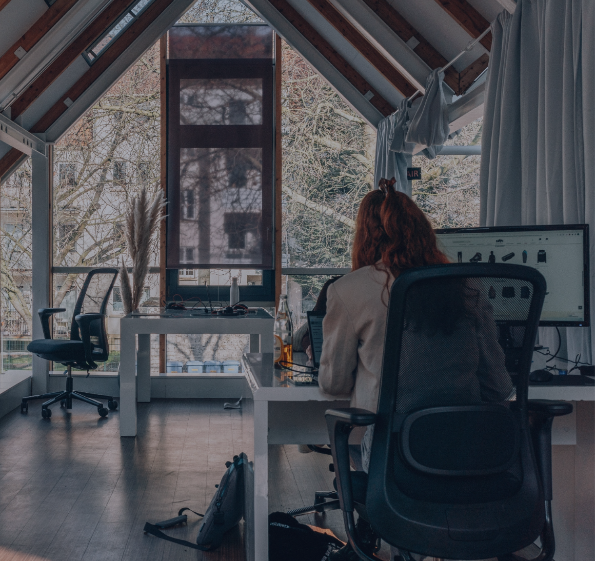 Person am Schreibtisch in einem hellen Büro mit großen Dachfenstern und Monitor
