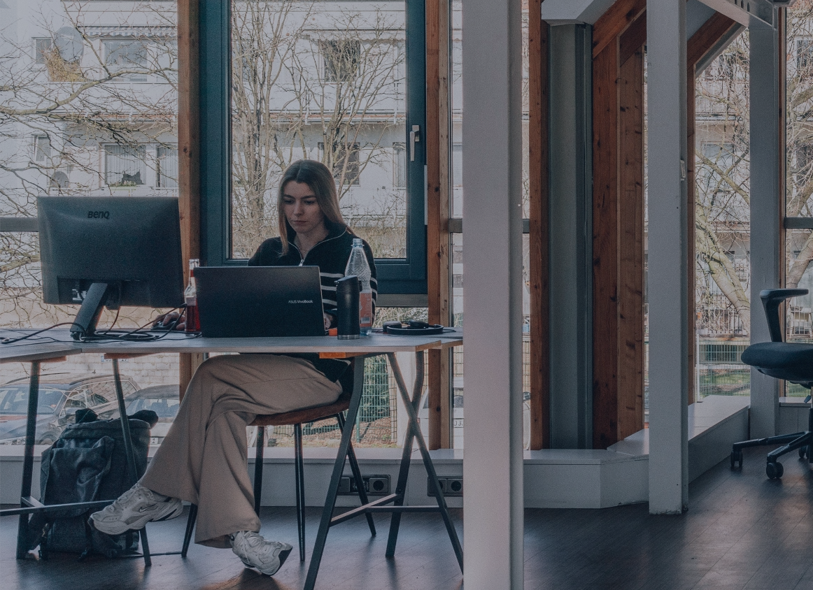 Person sitzt an einem Schreibtisch mit Laptop und Monitor vor großen Fenstern