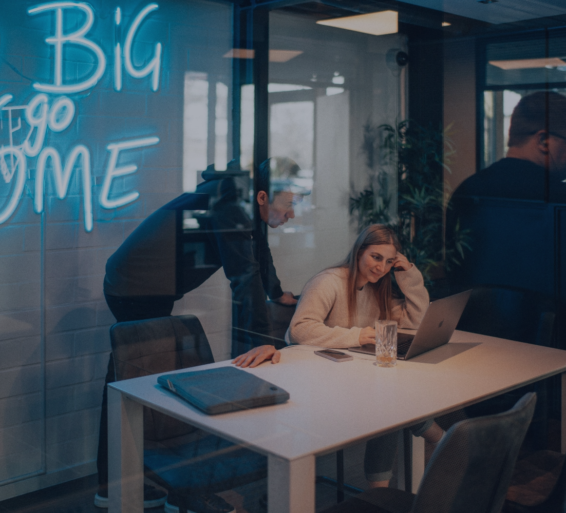 Zwei Personen arbeiten gemeinsam an einem Laptop in einem modernen Büro mit Neon-Schriftzug an der Wand