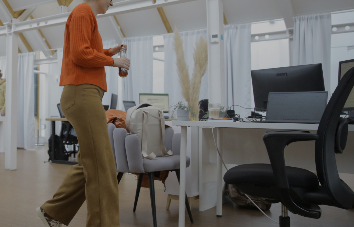 Person geht durch ein helles Büro mit Schreibtischen und Computern und hält eine Trinkflasche in der Hand.