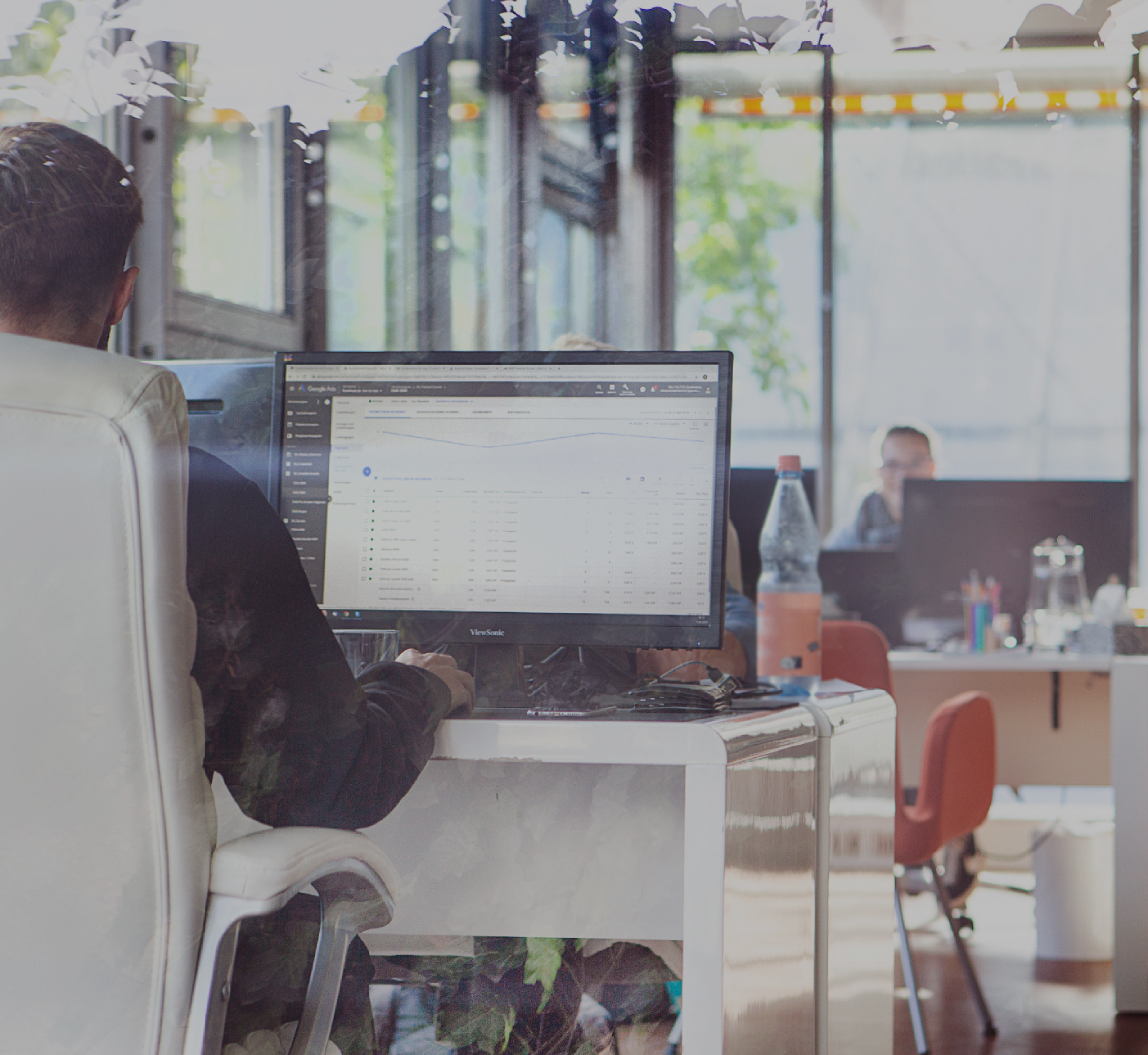 Heller Büroraum mit mehreren Arbeitsplätzen_ Im Vordergrund sitzt eine Person am Schreibtisch vor einem großen Monitor mit Tabellenansicht, im Hintergrund arbeiten weitere Personen an Laptops; eine Per