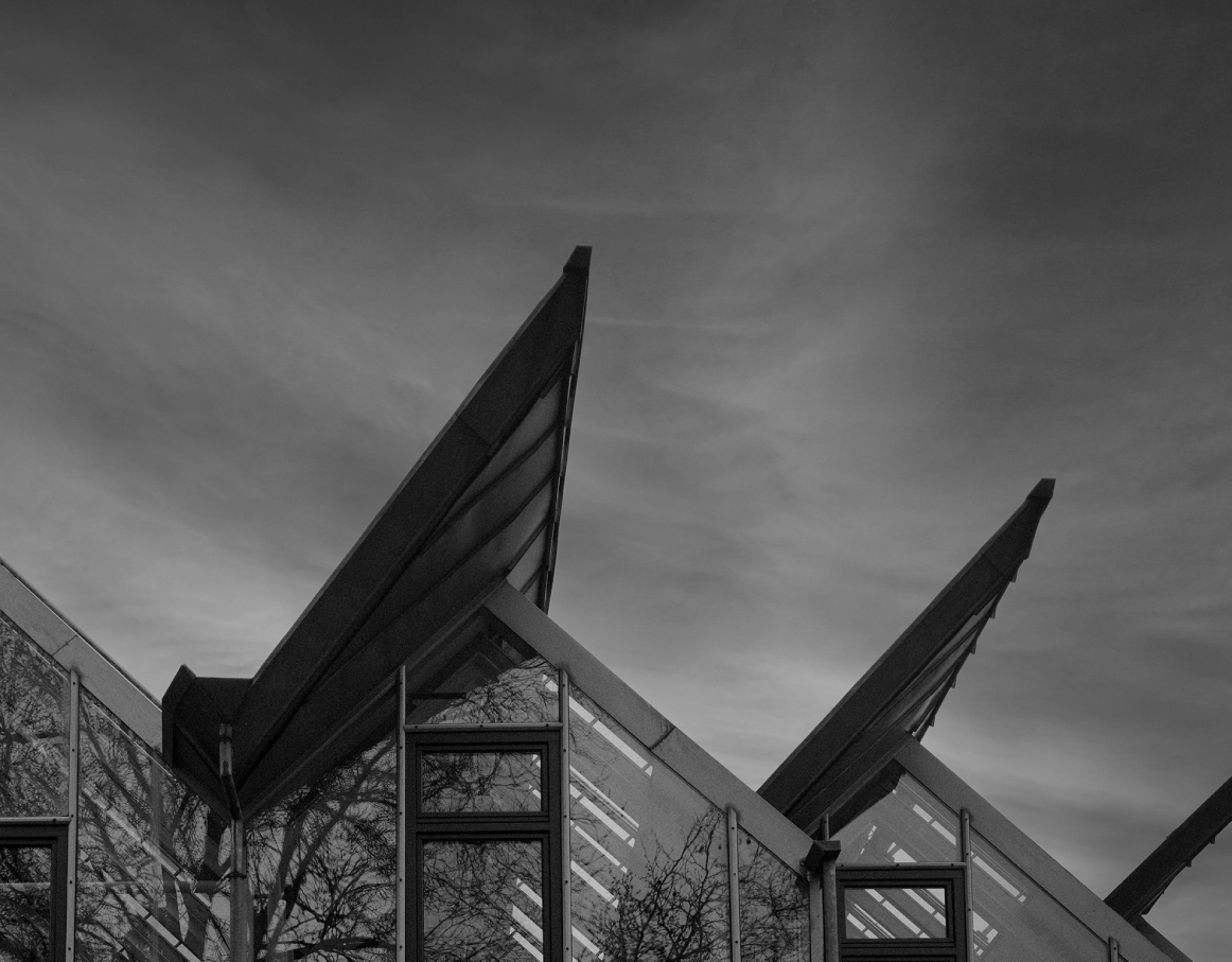 Schwarzweißaufnahme der Glasfassade eines modernen Bürogebäudes mit mehreren spitz zulaufenden, schräg auskragenden Dachkonstruktionen; Spiegelungen von kahlen Bäumen in den Fenstern und ein bewölkter