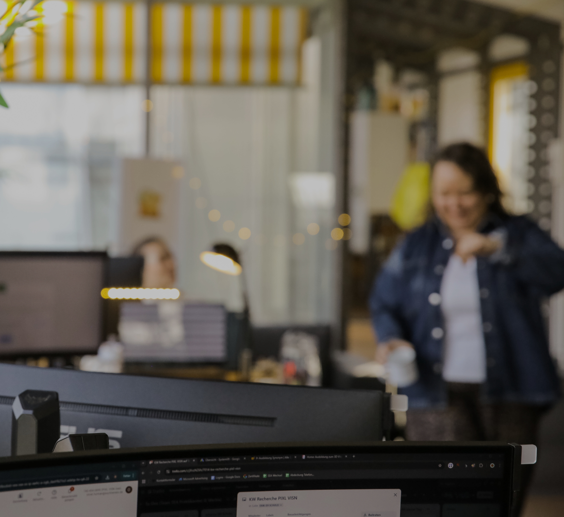 Unscharfe Büroszene mit Monitor im Vordergrund und einer Person, die im Hintergrund mit einer Tasse durch das Büro geht