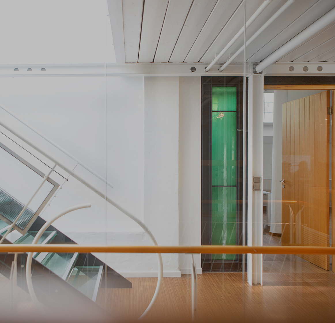 Heller Büroflur mit moderner Glas- und Metalltreppe, weißen Wänden und Holzboden; rechts ein offener Raum mit Holztür und grünem Glaselement, klare, minimalistische Architektur