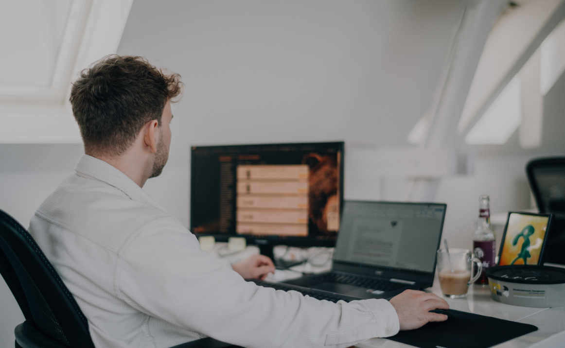 Person sitzt an einem Schreibtisch und arbeitet an einem Computer mit großem Monitor in einem hellen Büro, von hinten bzw. seitlich fotografiert