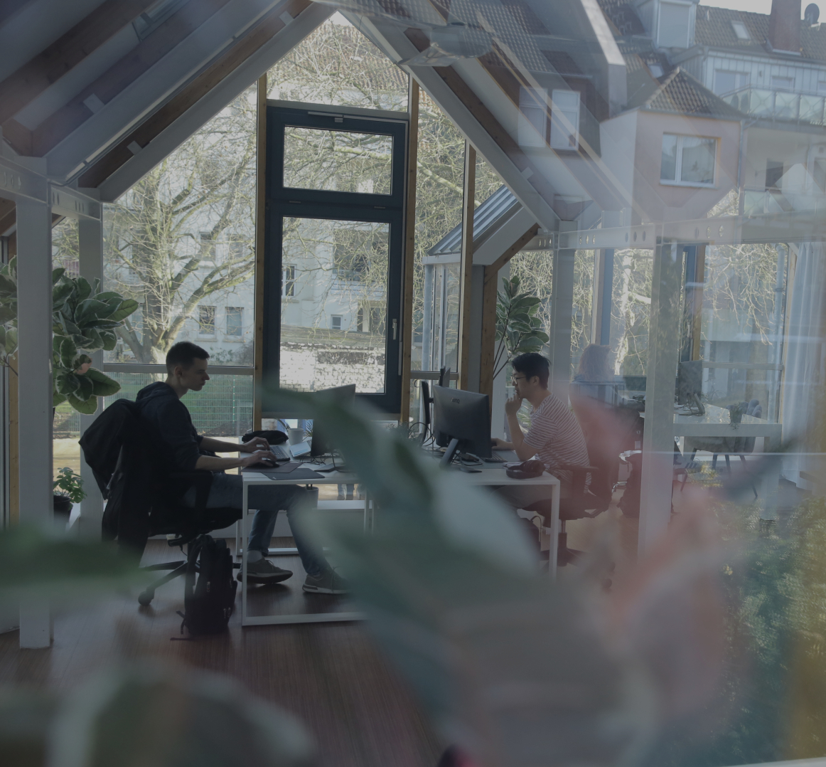 Zwei Personen arbeiten an Computern in einem hellen Büro mit großen Fenstern, Holzbalken und Pflanzen im Vordergrund.