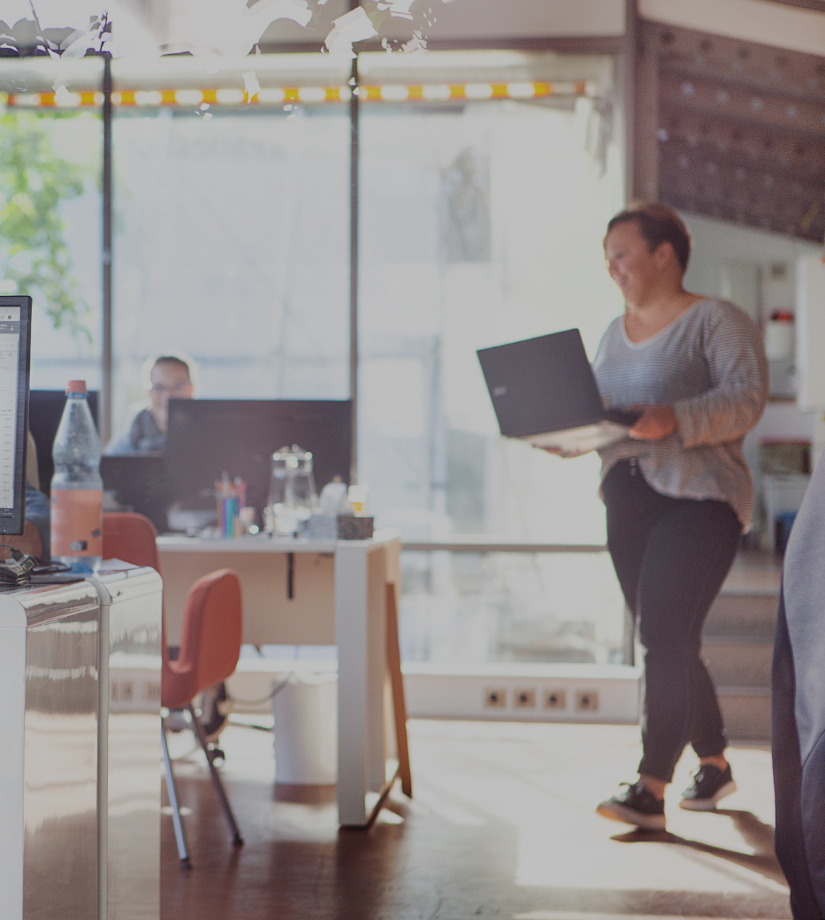 Heller Büroraum mit mehreren Arbeitsplätzen_ Im Vordergrund sitzt eine Person am Schreibtisch vor einem großen Monitor mit Tabellenansicht, im Hintergrund arbeiten weitere Personen an Laptops; eine Per