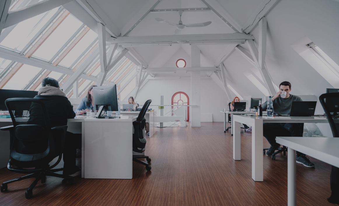 Helles Großraumbüro mit mehreren Personen an Schreibtischen unter einem Dach mit großen Fenstern.