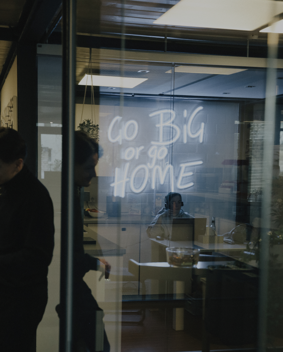 Blick durch eine Glaswand in einen Besprechungsraum mit leuchtender Neon-Schrift „Go Big or Go Home“ und arbeitenden Personen