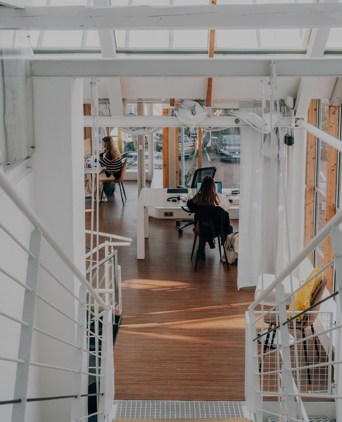 Blick von einer Treppe in ein helles, modernes Büro mit Holzfußboden, Glasfronten und zwei Personen, die an Schreibtischen arbeiten.
