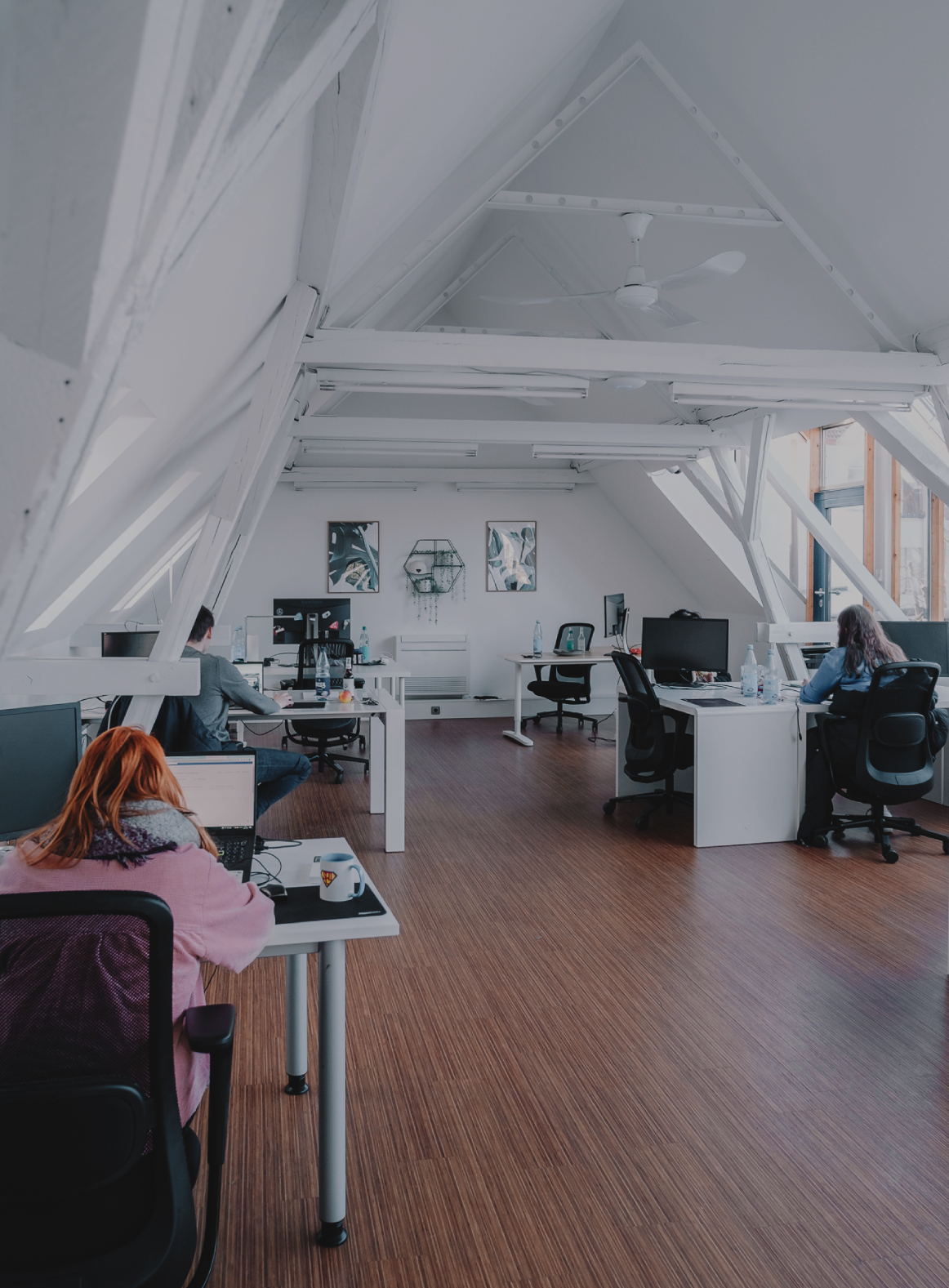Großzügiges, helles Büro mit schrägen Dachbalken, mehreren Arbeitsplätzen und Personen, die an Schreibtischen arbeiten