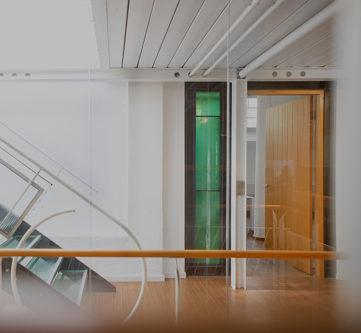 Heller Büroflur mit moderner Glas- und Metalltreppe, weißen Wänden und Holzboden; rechts ein offener Raum mit Holztür und grünem Glaselement, klare, minimalistische Architektur