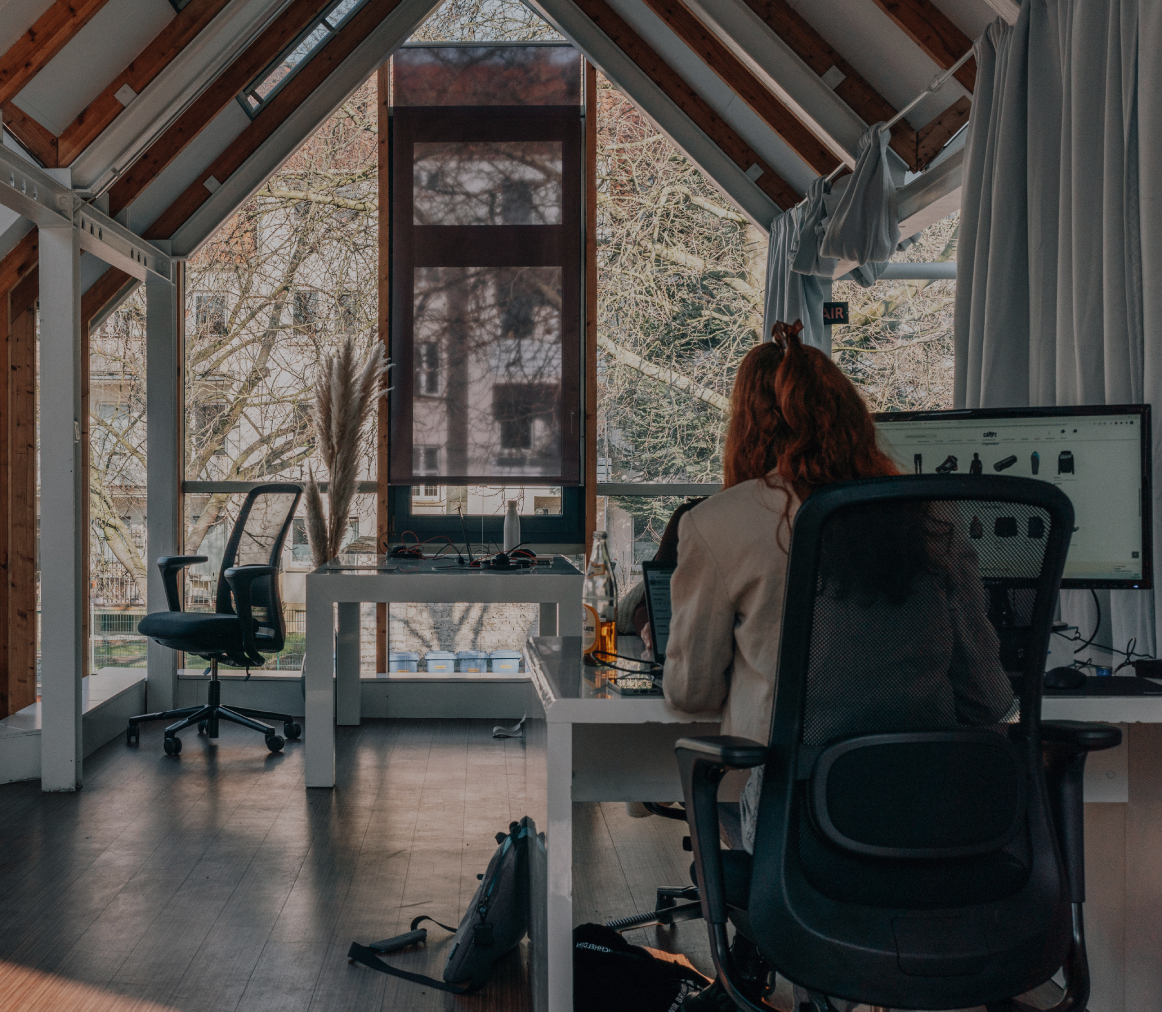 Zwei Personen arbeiten an separaten Schreibtischen in einem hellen Büro mit großen Fenstern, sichtbaren Holzbalken und Blick ins Grüne
