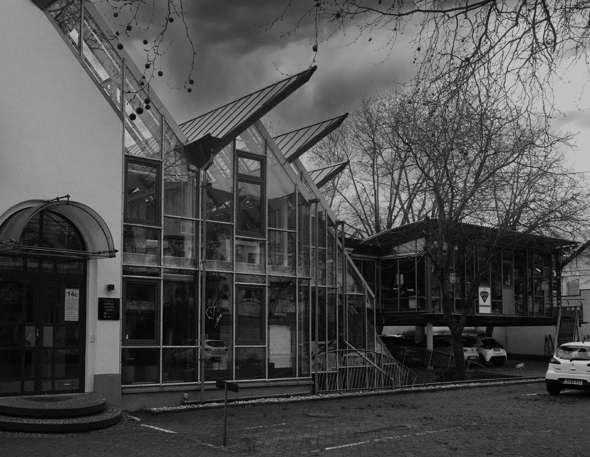 Schwarzweißaufnahme eines modernen Bürogebäudes mit großer Glasfassade und markanten, schräg auskragenden Dächern; davor Bäume ohne Blätter, ein gepflasterter Vorplatz und parkende Autos