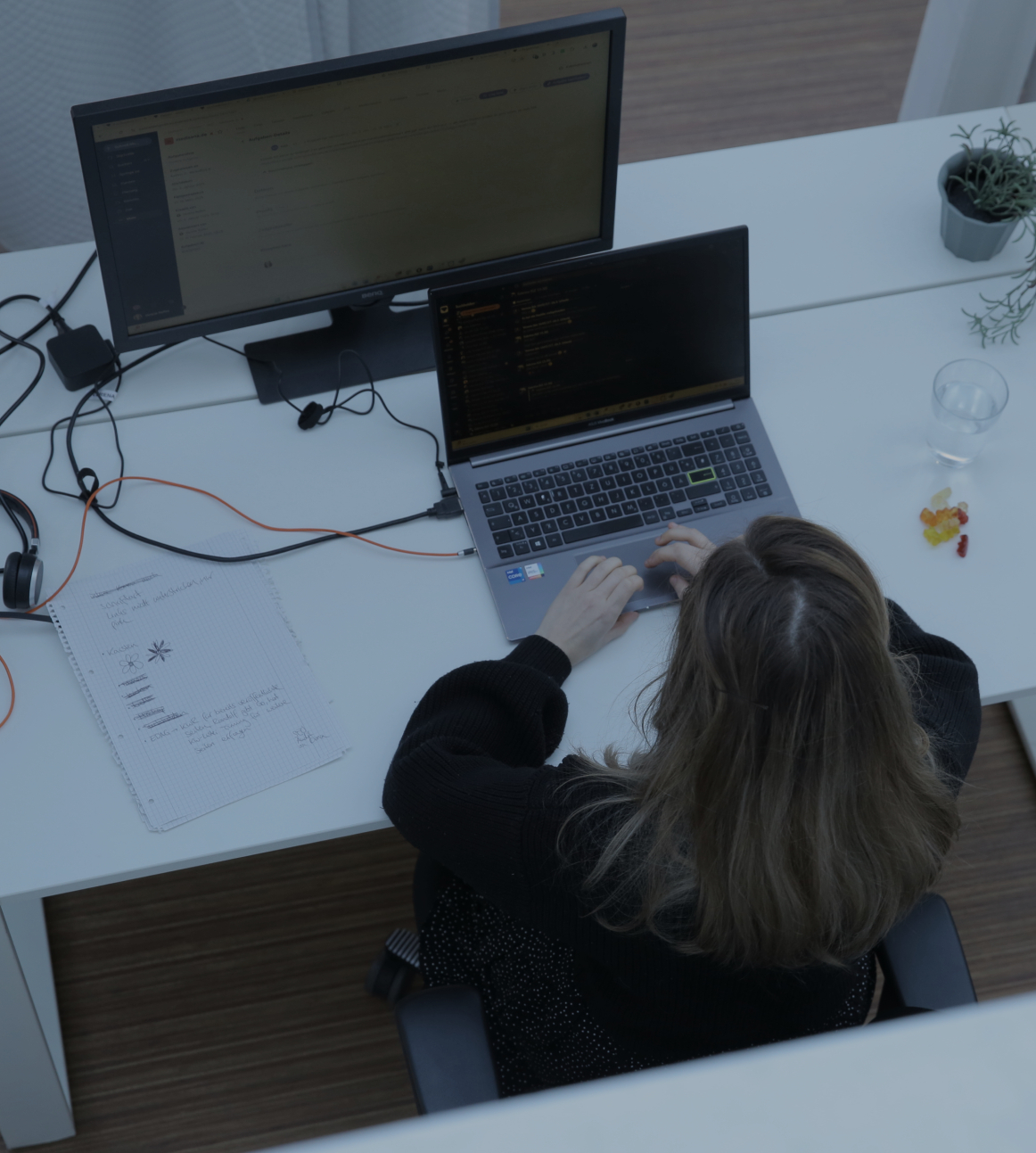 Person tippt an einem Laptop am Schreibtisch im Büro, aufgenommen aus der Vogelperspektive, mit externem Monitor und kleiner Pflanze