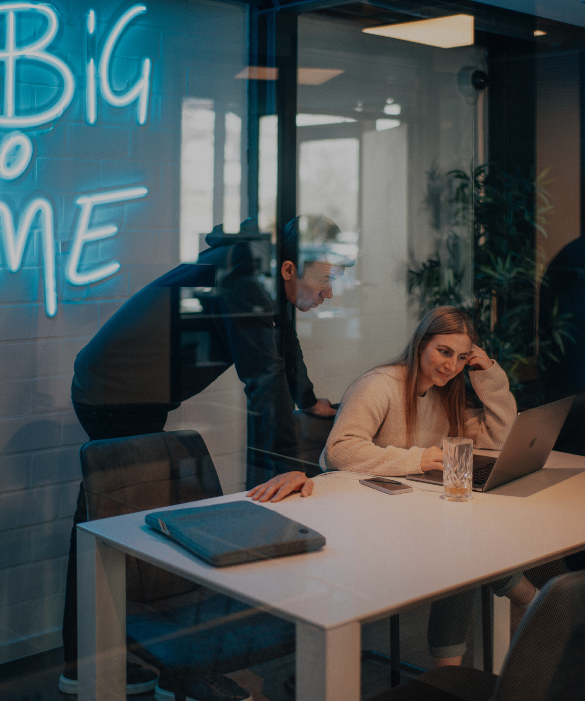 Zwei Personen arbeiten gemeinsam an einem Laptop in einem modernen Besprechungsraum, durch eine Glaswand fotografiert