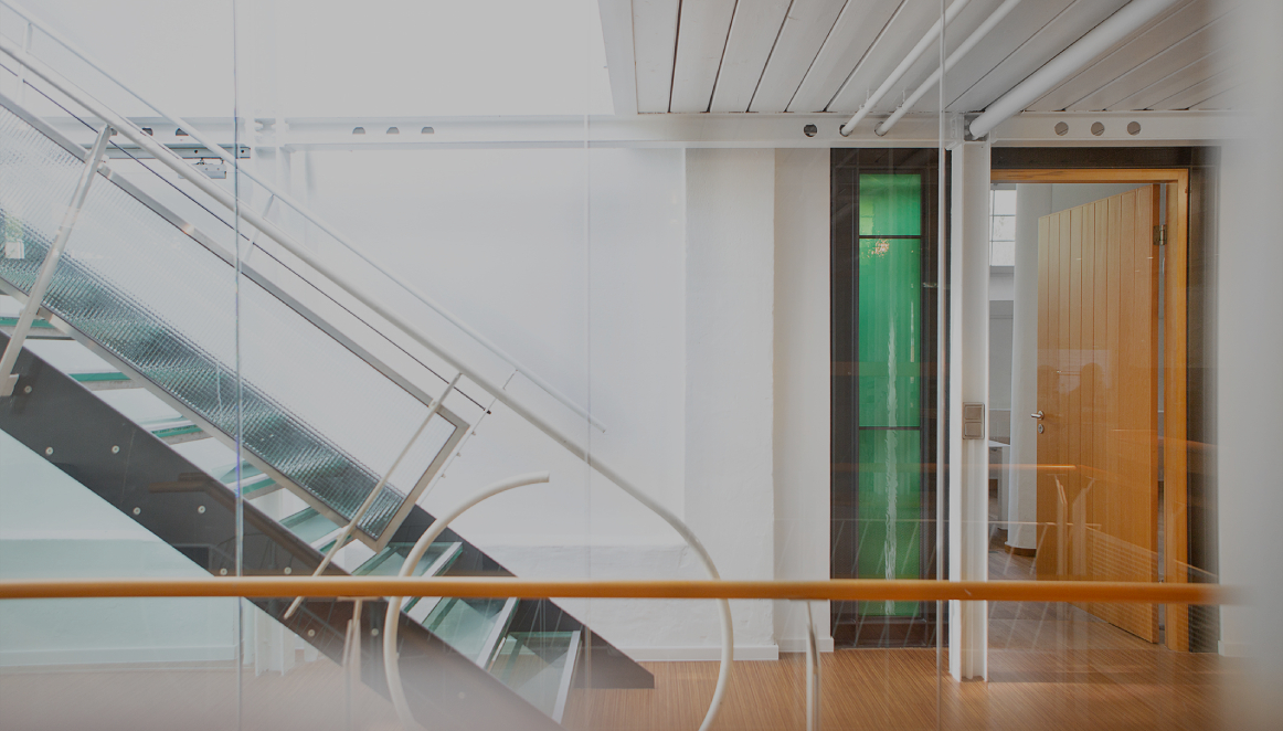 Heller Büroflur mit moderner Glas- und Metalltreppe, weißen Wänden und Holzboden; rechts ein offener Raum mit Holztür und grünem Glaselement, klare, minimalistische Architektur