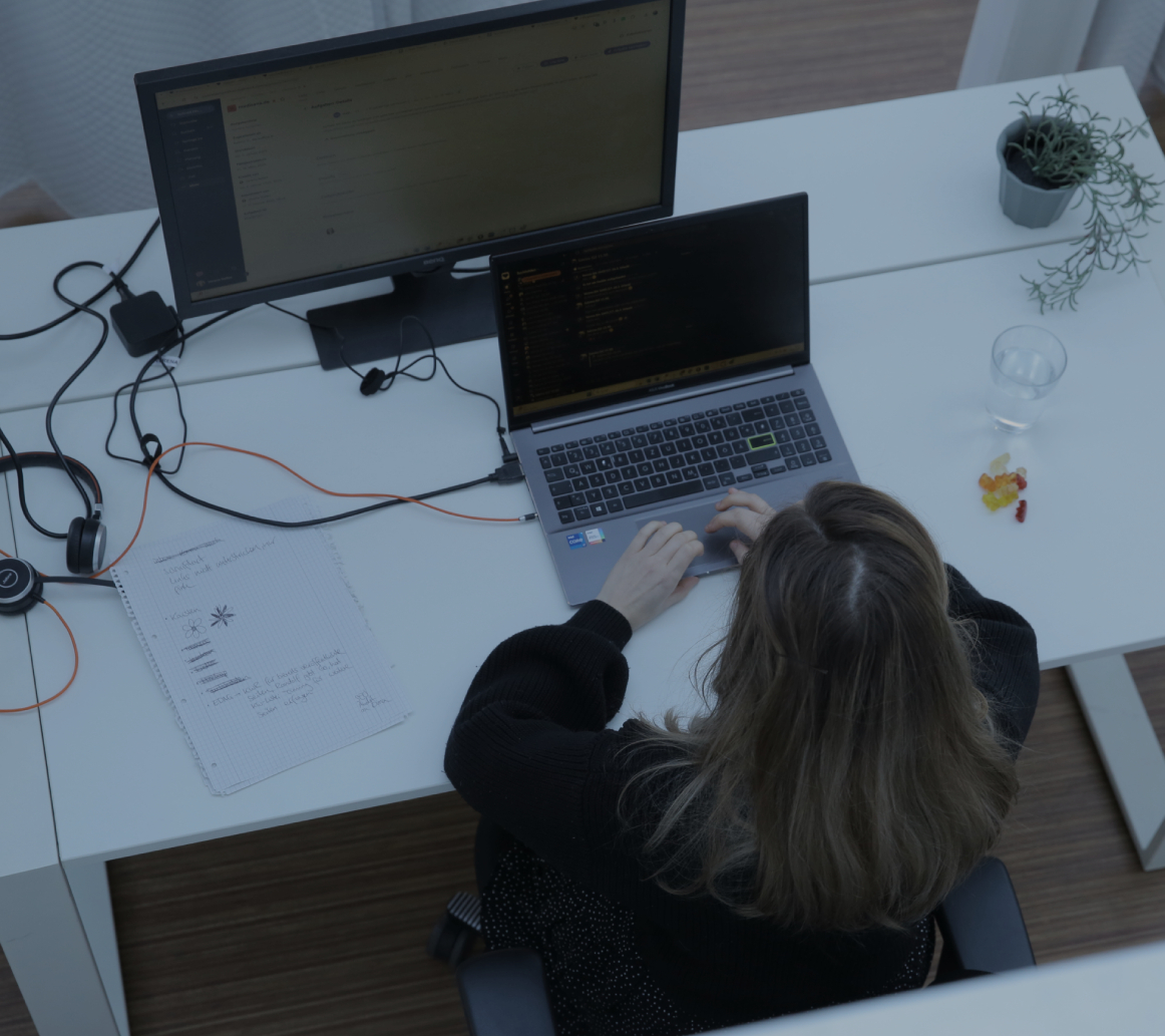 Person tippt an einem Laptop am Schreibtisch im Büro, aufgenommen aus der Vogelperspektive, mit externem Monitor und kleiner Pflanze
