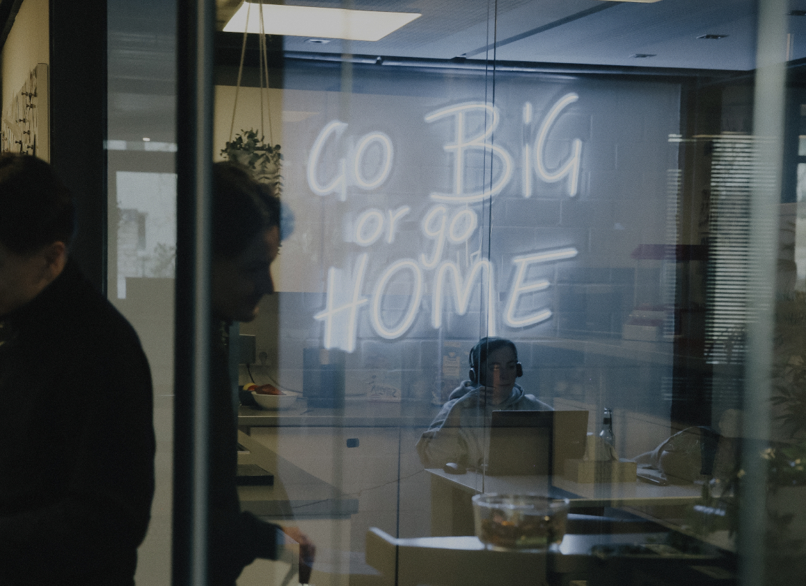 Blick durch eine Glaswand in einen Besprechungsraum mit leuchtender Neon-Schrift „Go Big or Go Home“ und arbeitenden Personen