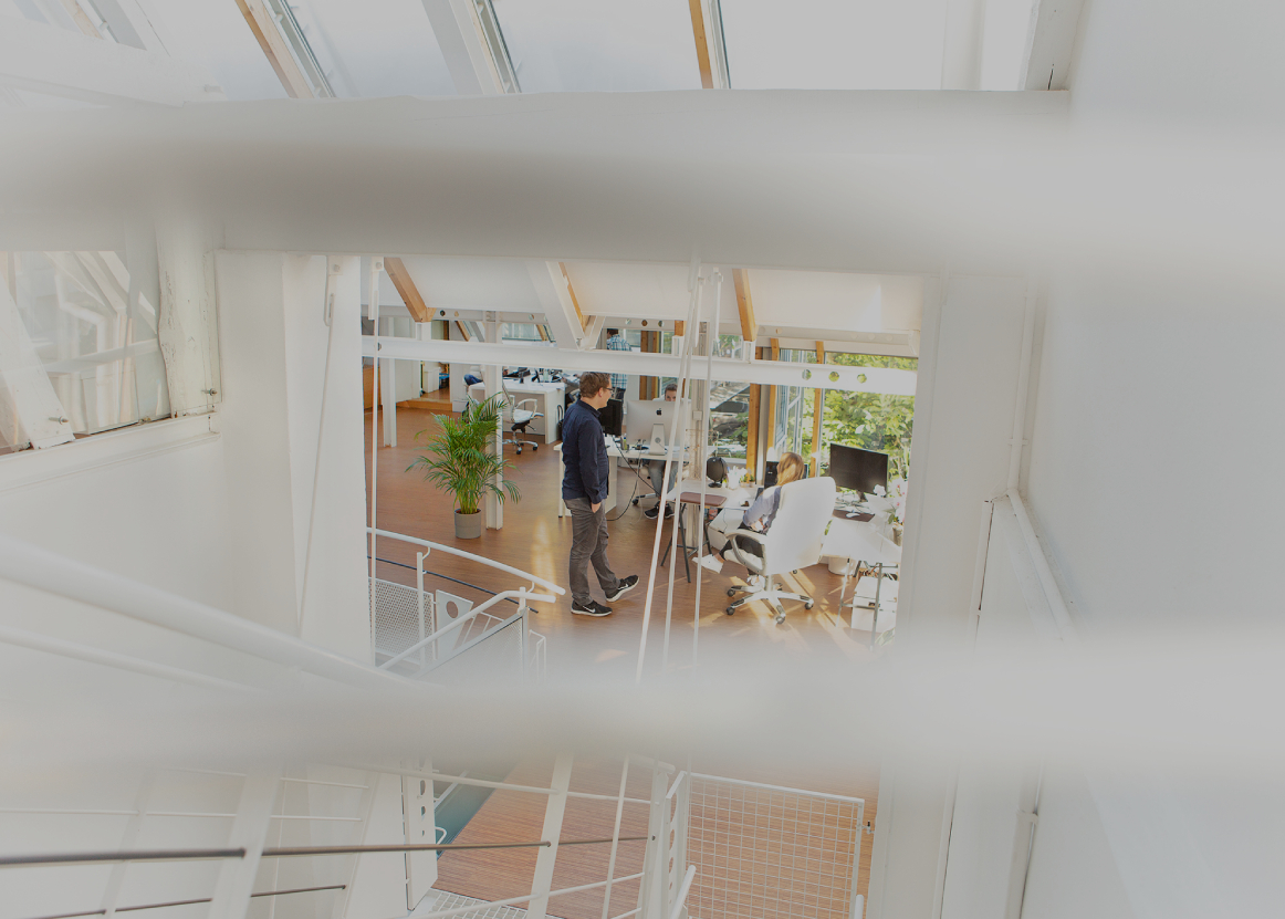 Offenes, lichtdurchflutetes Büro mit weißen Stahlträgern und Holzfußboden; mehrere Personen arbeiten an Schreibtischen, eine Person steht im Gespräch, große Fenster mit viel Tageslicht und Pflanzen im Raum